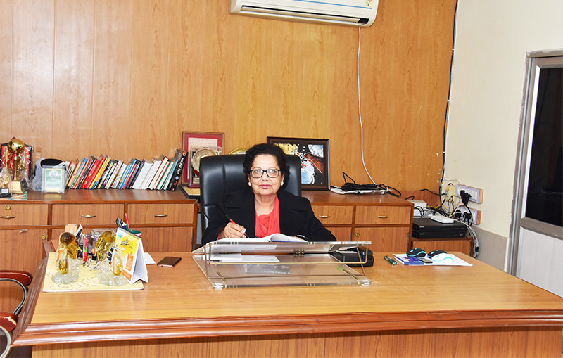 Woman in glasses and a black blazer writing at a large office desk.