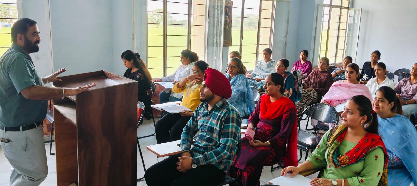 A lecturer in a dark green shirt speaks from a wooden lectern to about twenty seated students in a bright classroom, most of them women in salwar kameez.