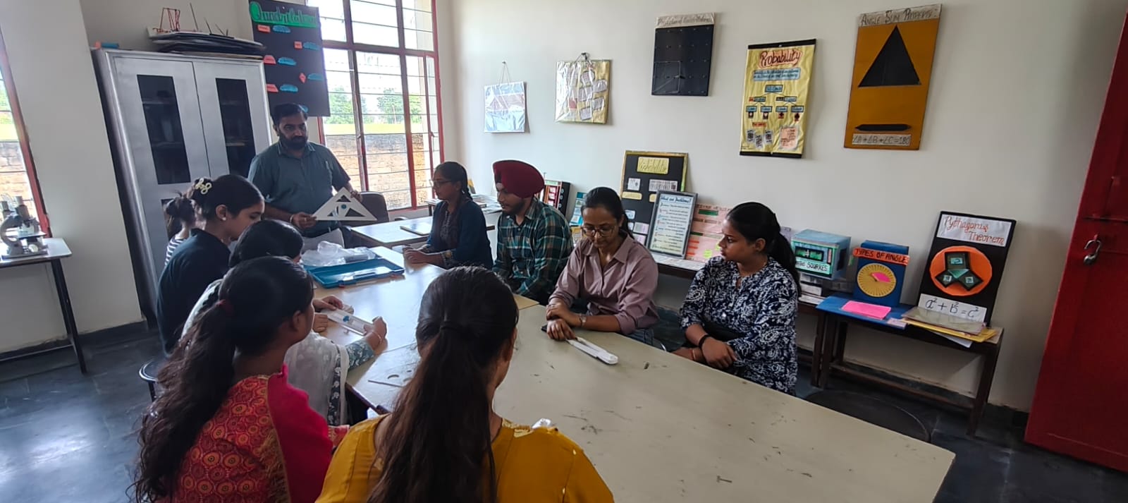 The mathematics laboratory: a lecturer holds up a card triangle to students seated around a long table, with charts on probability, the angle-sum property and Pythagoras' theorem on the walls.