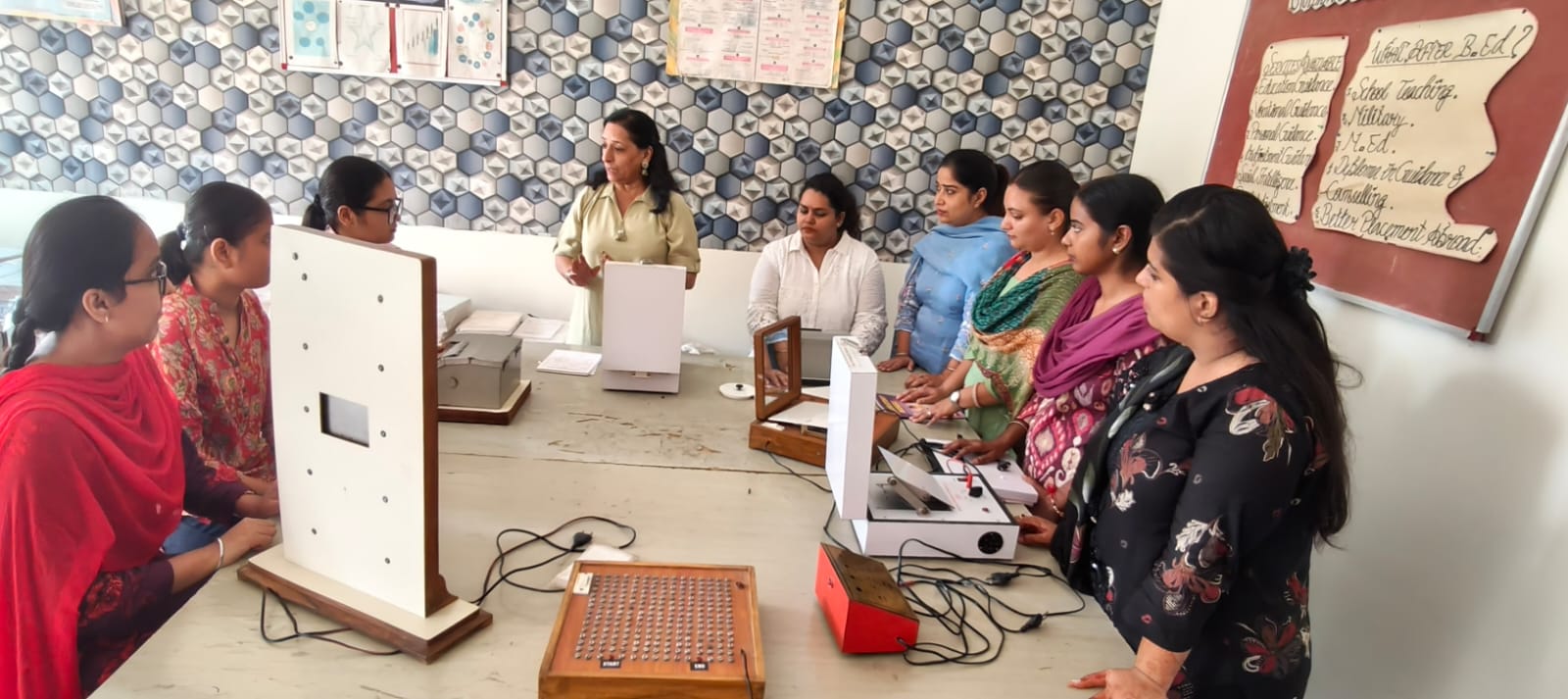 The psychology laboratory: a lecturer demonstrates testing apparatus on a table to students standing around it. A Guidance and Placement Cell board on the right lists careers open after a B.Ed.