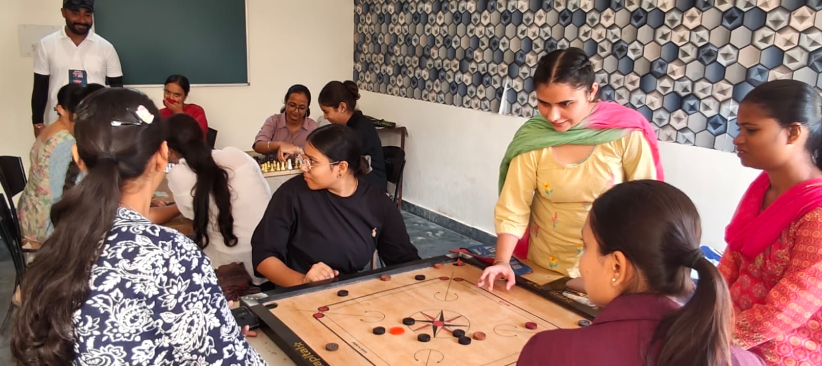 Students gathered around a carrom board mid-game, with a chess game in play at a table behind them.