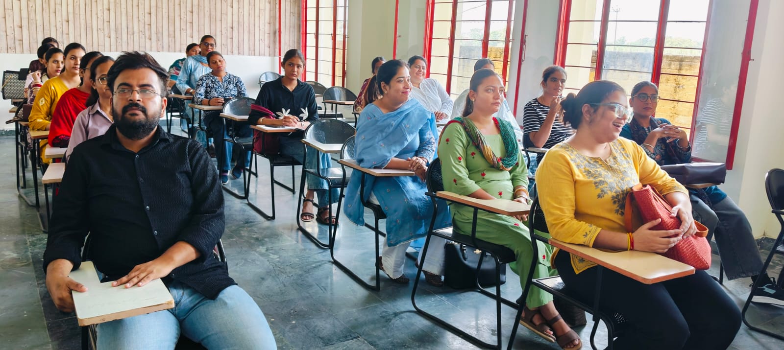 Students seated at tablet-arm chairs in a hall with red-framed windows, listening to a talk.