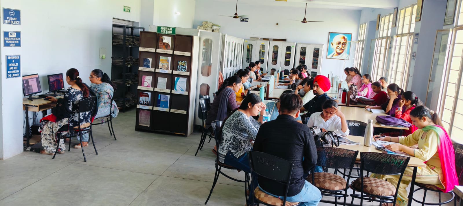 A wide view of the library: students read at long tables, a journal rack stands in the middle of the room, and signs on the left wall ask readers to keep silence.