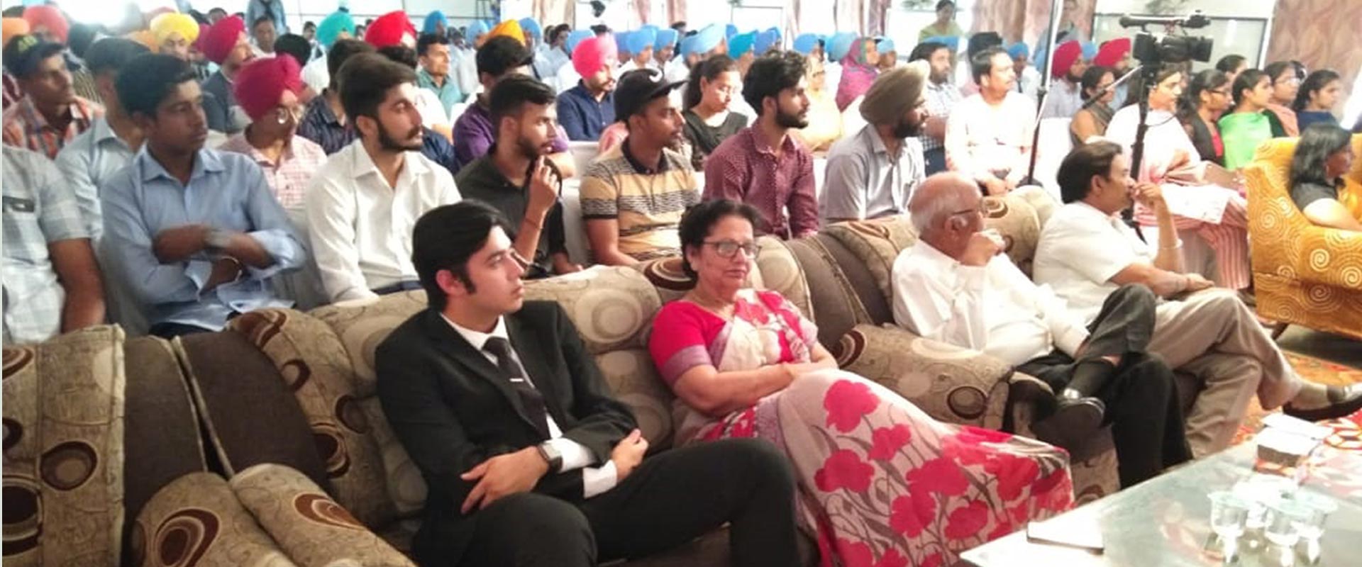 A crowded hall of seated people, many in turbans, facing forward as a camera films.