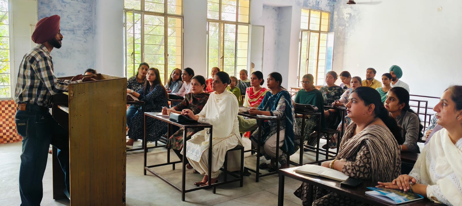 A lecturer in a maroon turban speaks from a lectern while students at bench desks look towards him, tall windows behind them.